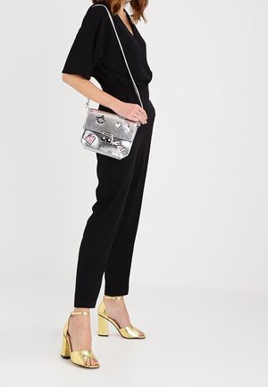 Žena v černém overal, zlatých sandálech na vysokém podpatku a s stříbrnou kabelkou přes rameno s dekorativními nášivkami.