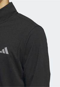 T-shirt de sport noir à manches longues avec col montant, montrant une partie d'un logo géométrique blanc imprimé sur la poitrine.