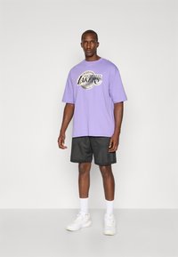 Tricou oversized violet cu grafică "Los Angeles Lakers", asociat cu șorturi din plasă negre. Adidași și șosete albe finalizează ținuta.