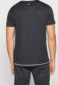 Svart atletisk skjorta med korta ärmar, gjord av strukturerad tyg; har vita accenter vid nederkanten och Fila-logotypen vid nackens bakdel.