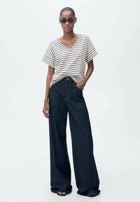 Randig, v-ringad t-shirt i krämfärg och svart, kombinerad med vida mörk denimjeans. Båda plaggen har en avslappnad passform och en avslappnad stil.