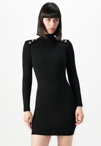 Donna con capelli scuri e lisci che indossa un vestito aderente nero a costine con maniche lunghe e bottoni dorati sulle spalle, in piedi contro uno sfondo bianco.