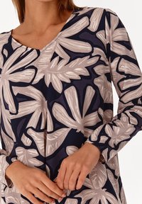 Pitkähihaisessa paidassa on V-pääntie, ja se on valmistettu kankaasta, jossa on tummansininen tausta ja vaaleanbeigejä kasvi-lehtikuvioita. Sileä pinta.