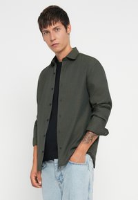 Młody mężczyzna z krótkimi brązowymi włosami, w otwartej ciemnozielonej koszuli zapinanej na guziki noszonej na czarnym t-shircie oraz jasnoniebieskich dżinsach, z tatuażem na lewym przedramieniu.