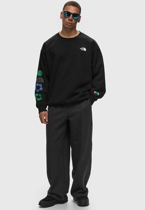 Sudadera negra de cuello redondo con el logotipo en el pecho y patrones coloridos en las mangas. Combinada con pantalones holgados negros y zapatillas negras.