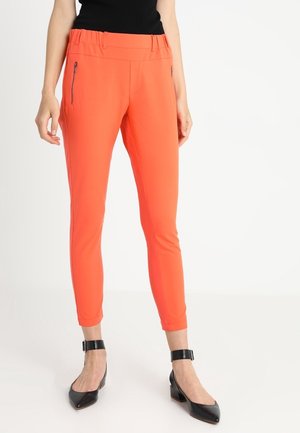 Schmal geschnittene, leuchtend orangefarbene Hose mit seitlichen Reißverschlusstaschen, kombiniert mit schwarzen spitz zulaufenden Pumps mit Knöchelriemen und einem schwarzen ärmellosen Oberteil.