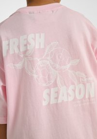 Ljusrosa kortärmad t-shirt med vit text "FRESH" och "SEASON" samt illustrationer av två fotbollar och löv.
