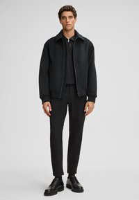 Czarna kurtka bombowiec z wyłogowym kołnierzem, zapinana na zamek, z bocznymi kieszeniami. Nosi ją osoba na czarnym topie, w połączeniu z dopasowanymi czarnymi spodniami i butami.