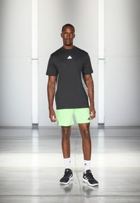 Zwart sport T-shirt met een wit logo, gecombineerd met neon groene shorts met witte accenten. Zwarte sportschoenen en witte sokken maken de outfit compleet.