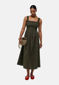 Robe midi sans manches vert olive avec un corsage froncé et de larges bretelles ; assortie à un sac rond en osier et des ballerines rouges.