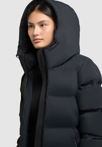 Černá péřová bunda s velkou kapucí, prošívaným designem a zipem v přední části. Obsahuje logo na zipu a hladkou strukturu.