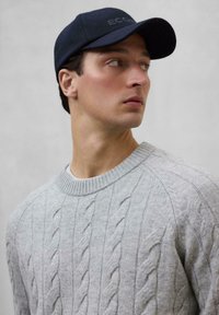 Suéter de punto trenzado gris con un escote redondo, superpuesto a una camisa blanca. Gorra azul marino con "ECC" bordado en la parte frontal.