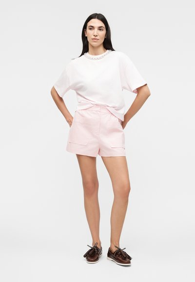 Γυναίκα με μακριά σκουρόχρωμα μαλλιά στέκεται με τα χέρια στη μέση, φοράει λευκό διακοσμημένο μπλουζάκι, ροζ ανοιχτό σορτς και σκούρα καφέ μοκασίνια με φούντες.