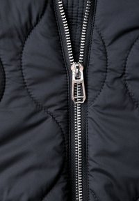 Gros plan sur une fermeture éclair en métal argenté partiellement fermée sur un tissu matelassé noir avec un motif de couture ondulée.