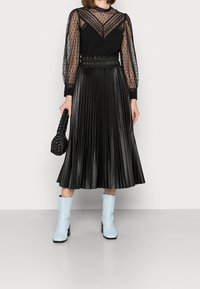 Schwarzer gefalteter Lederrock kombiniert mit einer durchsichtigen schwarzen Bluse, die mit Pünktchen versehen ist. Die Accessoires umfassen eine schwarze geflochtene Tasche und hellblaue Stiefel.