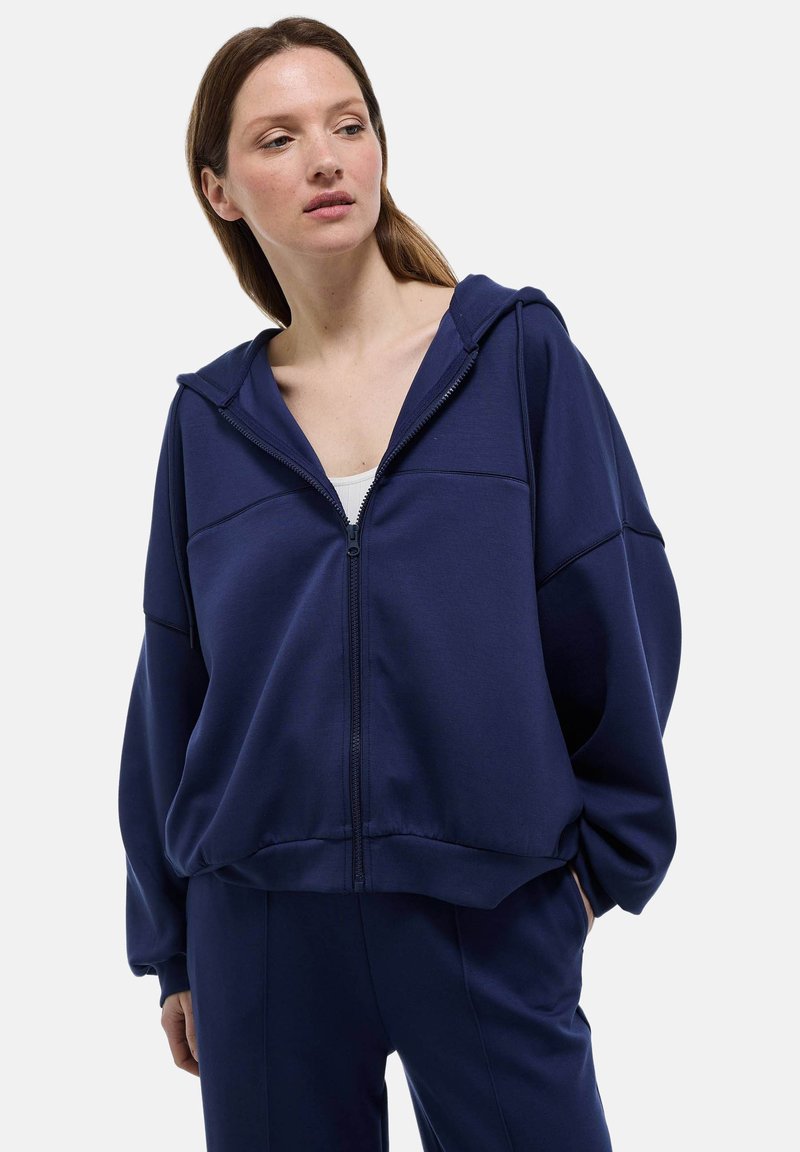 Sweat à capuche zippé bleu marine en tissu doux, avec une capuche doublée, des manches larges et une coupe décontractée, associé à un pantalon de jogging assorti.