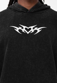Moletom preto com uma textura suave e capuz. Apresenta um design tribal de coração branco na parte do peito. Corte simples e estilo casual.
