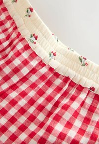 A piros és fehér vichy kockás anyag gyűrt derékrésszel rendelkezik, melyet egy világos virágminta és fodros vég díszít, hangsúlyozva a textúrát és a dizájnt.