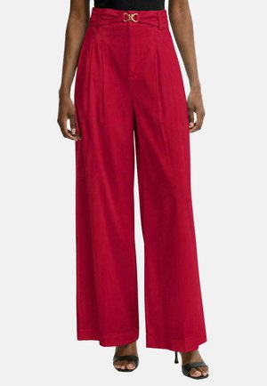 Donna che indossa pantaloni rossi a vita alta e gamba larga con cintura con fibbia dorata, abbinati a sandali neri aperti con tacco e top nero.