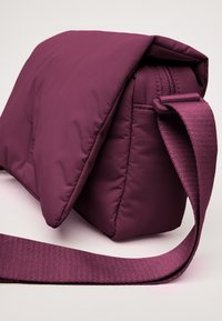 Borsa a tracolla in nylon viola con una soffice esterno imbottito, una parte superiore piatta con zip e una tracolla testurizzata. Forma rettangolare compatta.