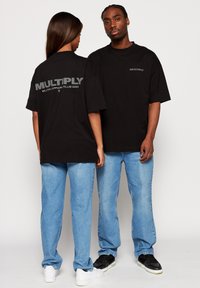 Mustat ylisuuret t-paidat, joissa on selässä harmaalla teksti "MULTIPLY APPAREL CLUB 2023". Mallit kokeilevat vaaleansinisiä farkkuja ja mustia tennareita.