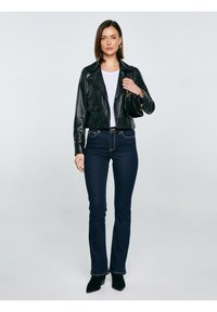 Žena u crnoj kožnoj jakni, bijeloj košulji, tamnoplavim trapericama, crnim čizmama i s crnom torbom na jednostavnoj svijetloj pozadini.