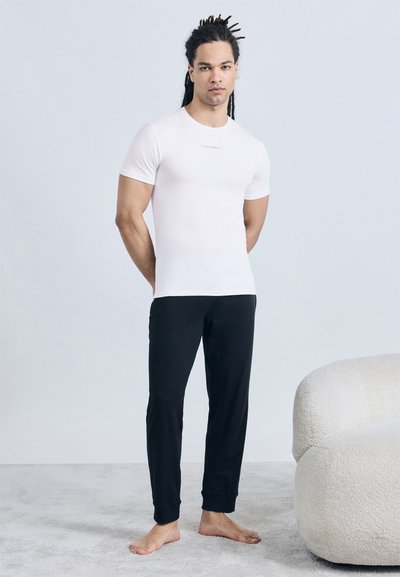 Άνδρας με ντραντλόκς στέκεται ξυπόλυτος πάνω σε μοκέτα, φορώντας εφαρμοστή λευκή μπλούζα και μαύρο παντελόνι φόρμας δίπλα σε καμπυλωτή λευκή πολυθρόνα.