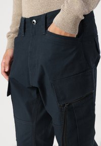 Pantalonii cargo de culoare albastru marin au multiple buzunare, un buzunar lateral cu fermoar și un închidere cu button la talie. Materialul are o textură fină.