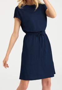 Vestido de manga corta azul marino con cintura ceñida y cordón. Tejido suave, corte relajado, con sutiles pliegues en los hombros.