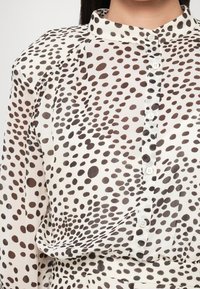 Fehér blúz fekete és barna absztrakt pöttyös mintával, magas gallérral és gombos előlappal, könnyű anyagból készült.