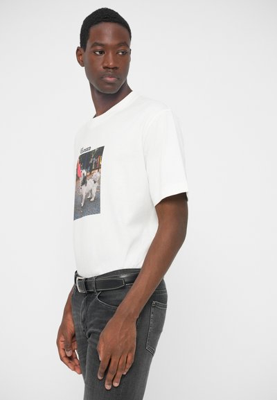 Tricou alb cu mânecă scurtă, având un print foto, decolteu rotund și o croială lejeră, purtat cu blugi gri închis și o curea neagră.