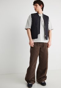 Νέος άνδρας που φοράει μαύρο γιλέκο με φερμουάρ πάνω από γκρι μπλουζάκι, φαρδιά καφέ παντελόνια και μαύρα αθλητικά παπούτσια, στέκεται μπροστά σε ένα απλό λευκό φόντο.