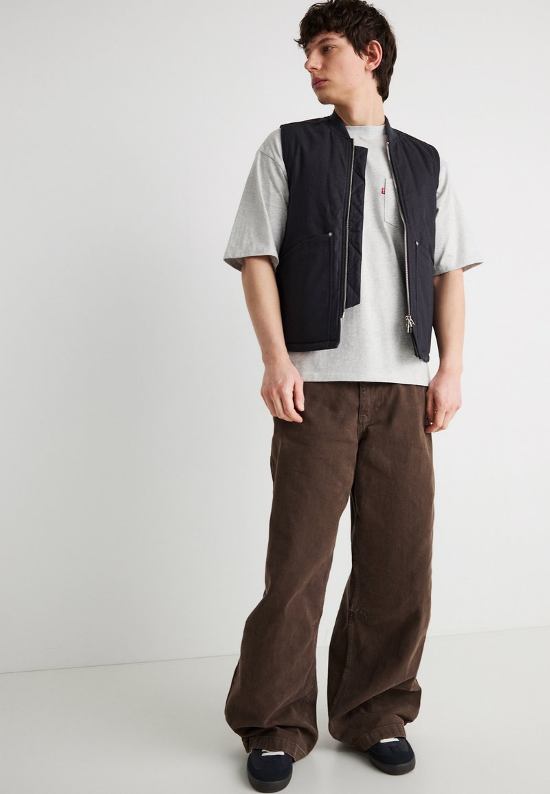 Giovane uomo che indossa un gilet nero con zip sopra una maglietta grigia, pantaloni marroni larghi e sneakers nere, in piedi davanti a uno sfondo bianco semplice.