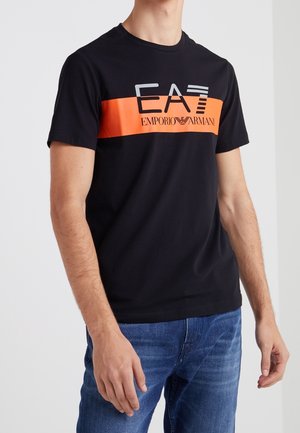 Uomo che indossa una T-shirt nera a maniche corte con una striscia arancione e il logo "EA7 Emporio Armani", abbinata a jeans blu, in piedi davanti a uno sfondo bianco.