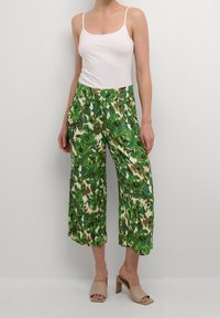 Pantaloni a palazzo verdi con motivo floreale, dalla vestibilità ampia e dotati di tasche laterali, realizzati in tessuto leggero. Abbinati a una canotta bianca e a tacchi beige.