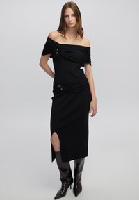 Zwarte gebreide jurk met een off-shoulder ontwerp, gerimpelde details en een zijsplit. Gepaard met zwarte leren laarzen tot aan de knie.