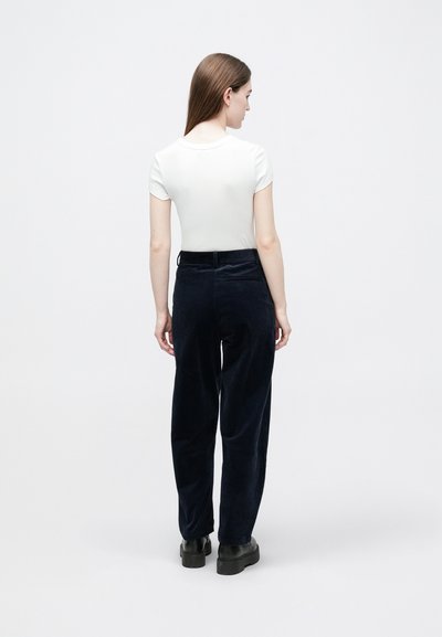 Sieviete ar gariem brūniem matiem, tērpusies baltā, piegulošā topā ar īsām piedurknēm, tumši zilu samta biksēs ar augstu vidukli un melnās platformas apavos, stāvot ar muguru pret skatītāju.