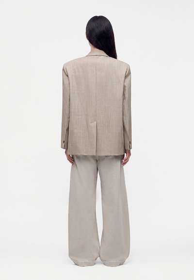 Άτομο με μακριά σκούρα μαλλιά φοράει μπεζ oversized σακάκι και παντελόνια με φαρδιά πόδια, στέκεται με την πλάτη προς ένα απλό λευκό φόντο.