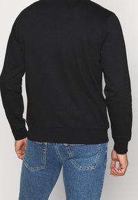 Felpa nera con maniche lunghe, polsini e orlo a coste, e una texture semplice e liscia. Indossata con jeans di denim blu con dettagli di cucitura.