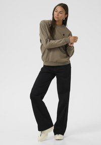 Pulover khaki cu guler rotund, având un mic logo cu un inimioară neagră, asortat cu pantaloni largi negri și adidași de culoare deschisă. Textură de țesătură netedă.