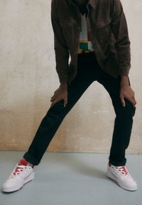 Braunes Kordhemd über einem mehrfarbigen, geometrisch bedruckten Hemd, kombiniert mit einer schwarzen Hose und weißen Sneakern mit roten Details.