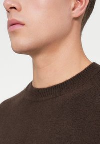 Närbild av en mans nedre ansikte och hals, iklädd en mörkbrun stickad tröja mot en enkel bakgrund.