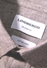 Camicia in cotone e lino marrone chiaro con una vestibilità rilassata. Etichette che indicano il marchio "Lindbergh" e il materiale. Texture visibile del tessuto.