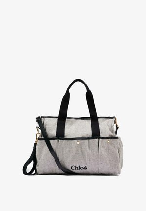 Bolso Chloé de tela beige con asas negras, bolsillos delanteros y una correa negra ajustable para el hombro sobre un fondo blanco.