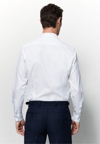 Witte katoenen langemouwen shirt met een puntige kraag, gezien vanuit de achterkant; heeft knopenmanchetten en een gladde textuur. Draagt met donkere broek.
