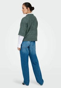 Groene textuurgebreide top met korte mouwen, laag over een witte blouse met lange mouwen, gecombineerd met losse blauwe denim jeans. Model is van achteren getoond.