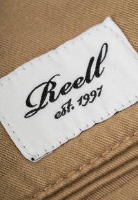 Beige fabric with a textured pattern, featuring a white sewn label that reads "Reell" in black cursive script with "est. 1997" below it.