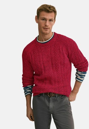 Młody mężczyzna o krótkich, jasnobrązowych włosach, ubrany w czerwony sweter w warkoczowy splot, nałożony na pasiastą koszulę z długim rękawem oraz szare spodnie z czarnym paskiem.