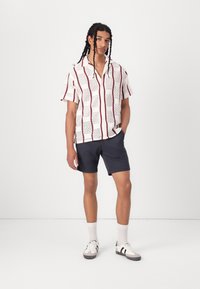 Korte mouwen shirt in het wit met een vierkant patroon en rode accenten, gecombineerd met donkere shorts en lichte sneakers, gedragen door een staande model.