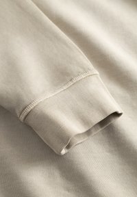 Κοντινό πλάνο ύφανσης μπεζ φούτερ, με υφή πλεκτού, μανσέτα με απλό τελείωμα και διακριτικές ραφές στις άκρες.